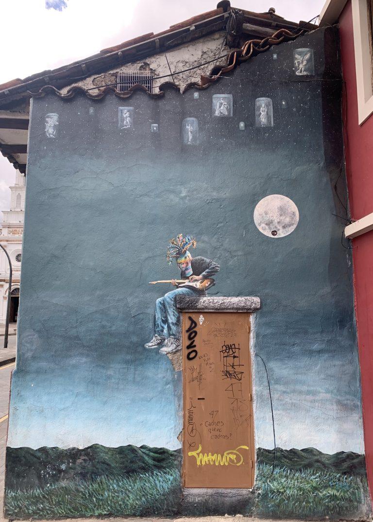 Street art of an electric guitarist wearing a traditional Cuenca headdress, playing on a building under a full moon